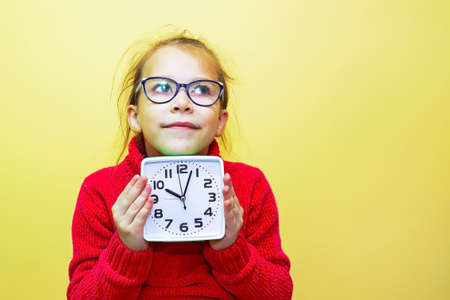 A child girl in a red sweater plays with an alarm clock, looks at the time and makes a gesture,...