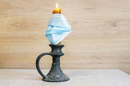 a lighted candle in a candlestick with a medical mask on it, which stands on the table against the...