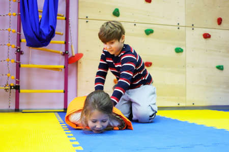 boy and girl children roll each other up in a thick blanket on mats controlling a small space for...