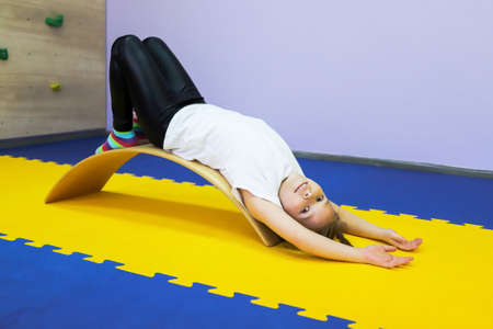 a little girl in a white T-shirt lies on a medical balance beam in a rehabilitation center and undergoes course of treatmentの写真素材