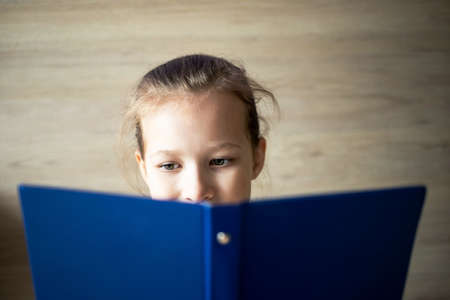 little girl child reads a blue book and looks out from behind it, eyes and a smile are visible, school tomorrow.の写真素材
