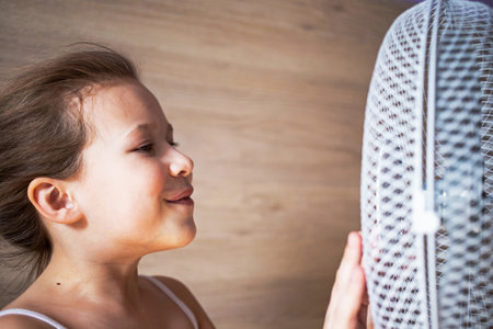 a little girl looks closely at the fan, touches the air with her hands, enjoys the flow of air in the heat.の写真素材