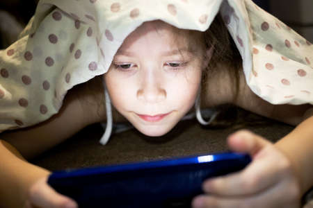 Cute little girl hiding under a blanket and watching a cartoon about magical space on the phone.の写真素材