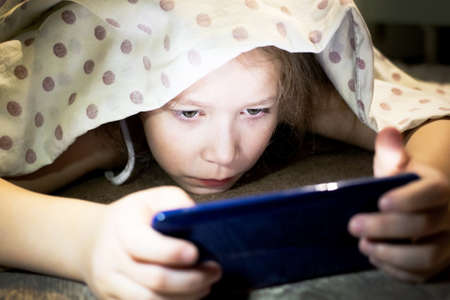 Cute little girl hiding under a blanket and watching a cartoon about magical space on the phone.の写真素材