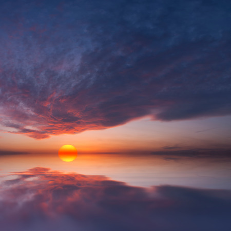 Sunset with big red sun and beautiful clouds reflected in waterの写真素材