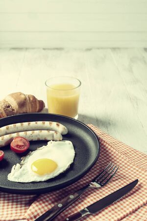 Fried sausage with eggs and vegetables on pan. Delicious breakfast with orange juise and croissant on wooden rustic boards, food background. Vintage stylization, retro film filterの写真素材