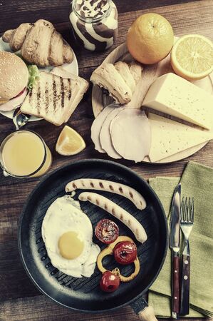 Fried sausage with eggs and vegetables on pan. Delicious breakfast with orange juise and croissant on wooden rustic boards, food background. Vintage stylization, retro film filterの写真素材