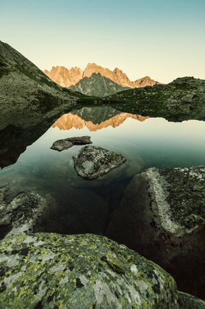 Sunlit mountain peaks are beautifully reflected in the water of a small lake, calm water and blue sky. Retro film filter, vintage stylizationの写真素材