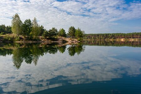 Summer landscape with forest on coastline and clouds reflection in water of lakeの写真素材