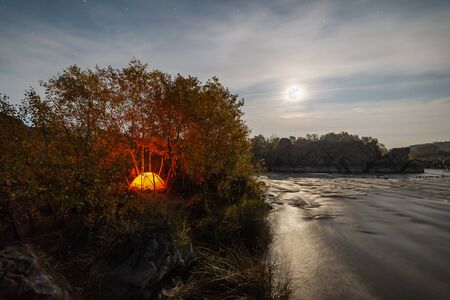 Night landscape with tent on shore of river and full moon and stars in sky. Travel card, long exposureの写真素材