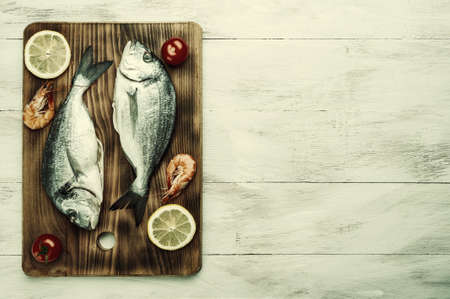 Fresh dorado fishes and shrimps on bord prepared to cooking, vegetables and fresh sea food. Cooking background, flat lay. Vintage stylization, retro film filterの写真素材