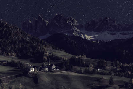 Rural landscape with church in famous village Santa Maddalena and Dolomites mountain peaks at night with stars and moon light. Vintage stylization, retro film filterの写真素材