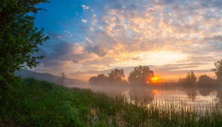 Summer landscape with river and majestic foggy sunrise with bright clouds in skyの写真素材