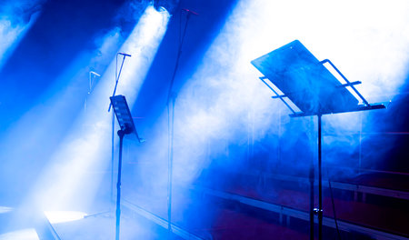 foggy stage with a blue light illuminating the area. Several microphones and stands set up for use during performanceの写真素材