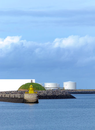North Mole Head yellow lighthouse and green park in the port of Reykjavik Iceland seawater against industrial constructions on sunny summer day under cloudy blue skyの写真素材