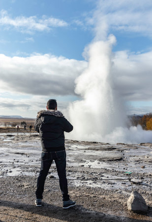 Back view of anonymous male traveler looking away while standing on rocky volcanic terrain with water in daylight and admiring gushing out white steam from natural geyser spring. Smidur Geysir. The Golden Circle, Icelandの写真素材