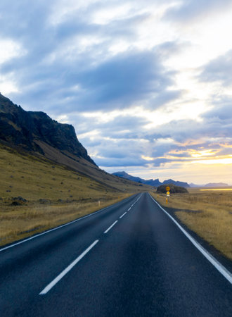 A long road with no cars on it and a mountain in the background. The sky is cloudy and the sun is settingの写真素材