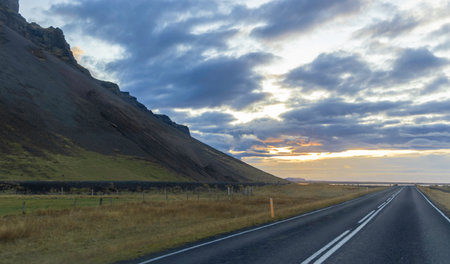 A road with a hill in the background and a cloudy sky. The sky is orange and the sun is settingの写真素材