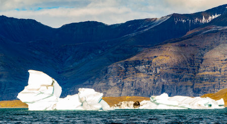 A large ice block is floating in the ocean. The ice block is surrounded by water and there are rocky mountains in the backgroundの写真素材