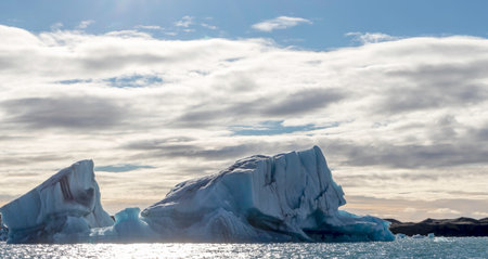 A large ice block is floating in the ocean. The sky is cloudy and the sun is shining through the...
