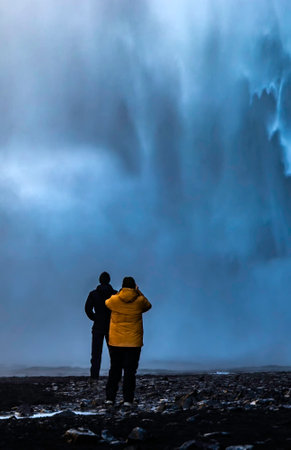 A man and woman stand in front of a waterfall, taking a photo. Scene is serene and peaceful, as the couple enjoys the beauty of natureの写真素材