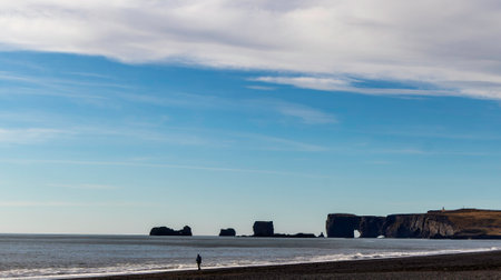 A person is walking on a beach with a view of the stone arches of Dyrholaey, Iceland. The sky is clear and blue, and the beach is emptyの写真素材