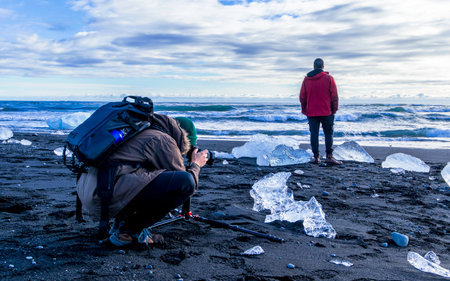 A man crouches down to take a picture of a block of ice washed up on the beach by the tide. The beach is covered in iceの写真素材