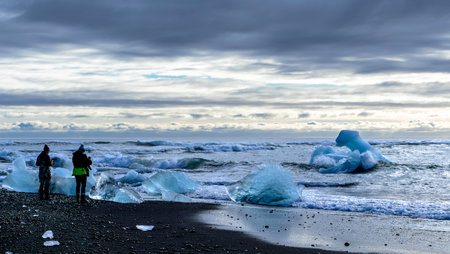 Two photographers are standing on a beach looking out at the ocean. The sky is cloudy and the water...