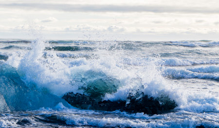 The ocean is rough and the waves are crashing against an iceberg. The water is blue and white. The sky is cloudy and the sun is not visibleの写真素材