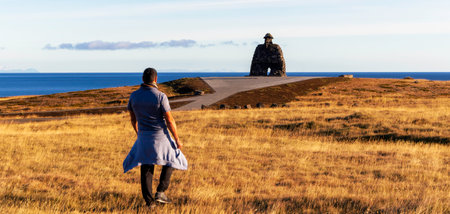 A man is walking on a field towards a statue of a man in the background. The sky is blue and the grass is yellow. Bardur Snaefellsas statueの写真素材