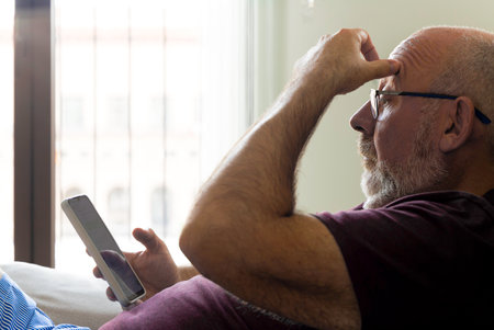 A man is sitting on a couch with a cell phone in his hand. He is looking at the phone with a puzzled...