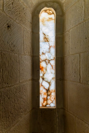 A narrow window with a stone frame and a stone arch. The window is lit by sunlight. The light shines...