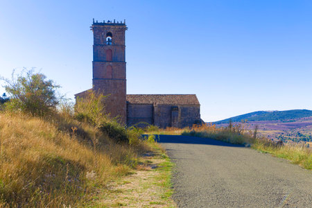 A small church with a bell tower sits on a dirt road. The road is surrounded by a dry, grassy hillsideの写真素材
