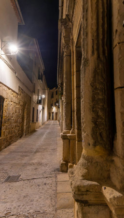 Streets of Baeza, Spain, by night. A narrow alleyway with a stone wall on the side. The street is lit up by street lights and the shadows of the buildings create a mysterious atmosphereの写真素材