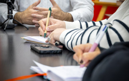 Two women are taking notes in their notebooks while a third person speaks. None of the people are...