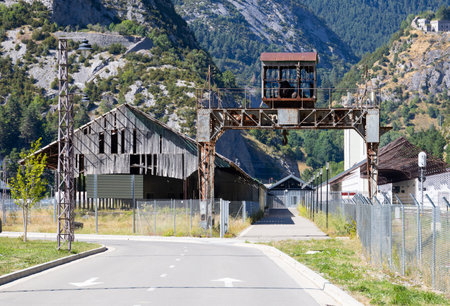 Wide shot of a rustic, abandoned railway station at the base of a steep mountain. Features a rusty metal structure and old wooden sheds. Industry, decay, historyの写真素材