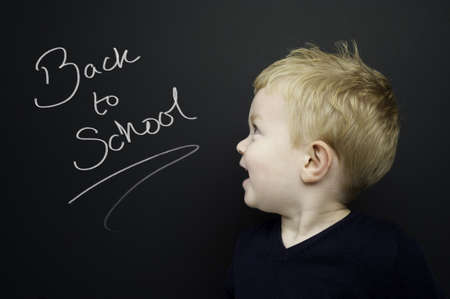Smart young boy wearing a navy blue jumper stood infront of a blackboard with Back to School written...