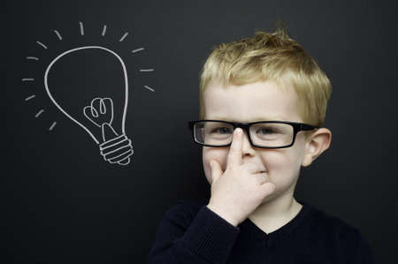 Smart young boy wearing a navy blue jumper and glasses stood infront of a blackboard with a drawn on chalk light bulbの写真素材