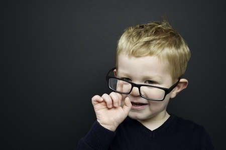 Smart young boy wearing a navy blue jumper and glasses stood infront of a blackboardの写真素材