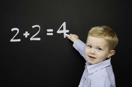 Smart young boy wearing a blue striped shirt stood writing a math sum on a blackboardの写真素材