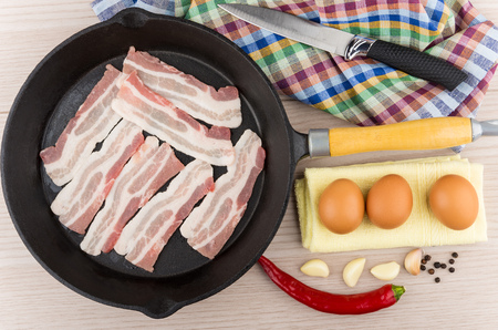 Pieces of raw bacon in cast iron frying pan, eggs, garlic, chili peppers and kitchen knife on wooden table, top viewの写真素材