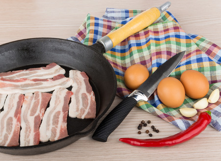 Pieces of raw bacon in cast iron frying pan, eggs, garlic, chili peppers and kitchen knife on towelの写真素材