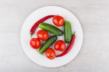 Tomatoes, cucumbers and red hot chili peppers in white glass plate on wooden table. Top viewの写真素材
