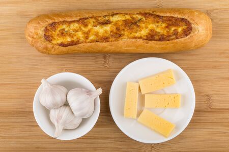 Baguette, garlic in bowl and pieces of cheese in saucer on wooden table. Top viewの写真素材