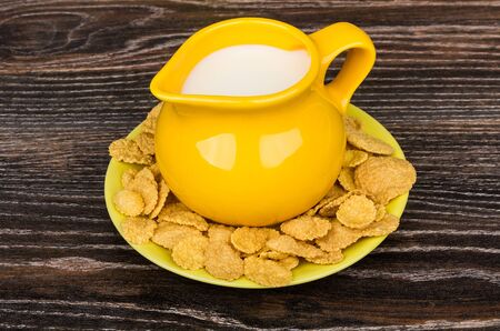 Yellow jug milk in saucer with corn flakes on dark table