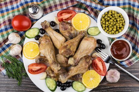 Heap of fried chicken legs with vegetable, black olives and lemon in dish on checkered towel. Top viewの写真素材