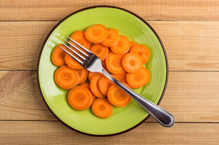 Slices of raw carrot in green plate and fork on wooden table. Top viewの写真素材