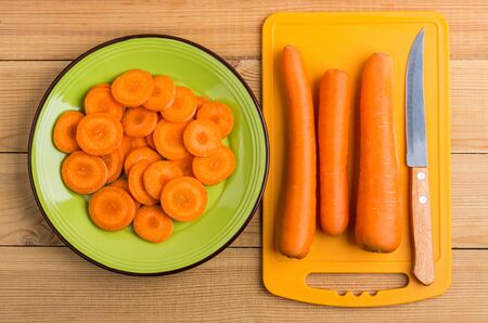 Slices of raw carrot in green plate, cutting board and knife on wooden table. Top viewの写真素材