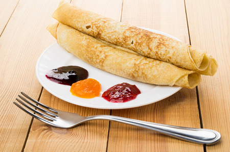 Rolled up pancakes in plate, peach, blueberry, raspberry jam and fork on wooden tableの写真素材