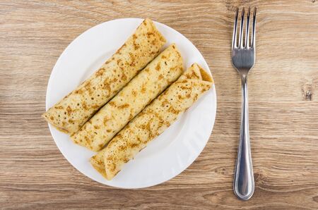 Three pancakes with stuffing in white plate and fork on wooden table. Top view